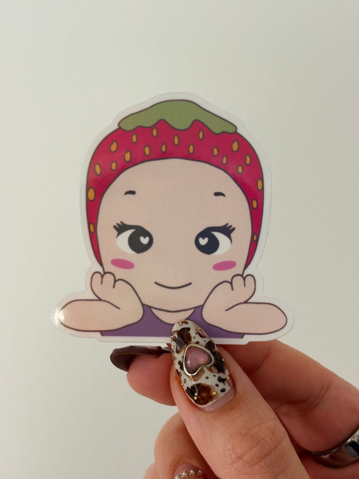 A die-cut sticker featuring a cute cartoon character with a pink strawberry-patterned headband and purple top, held by a hand with leopard-print nail art against a plain light background.