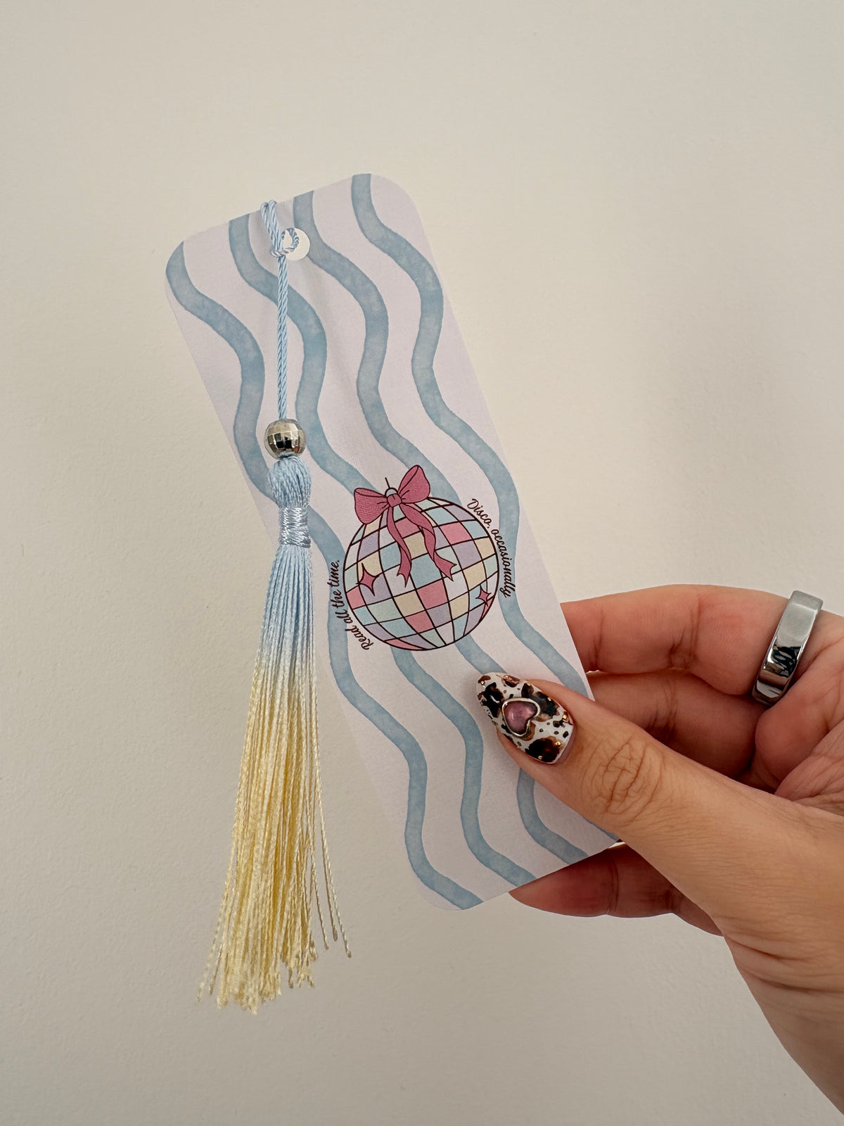 A hand holding a decorative paper bookmark featuring light blue wavy stripes, a pink-and-white disco ball illustration with a bow, and a light blue and cream tassel.