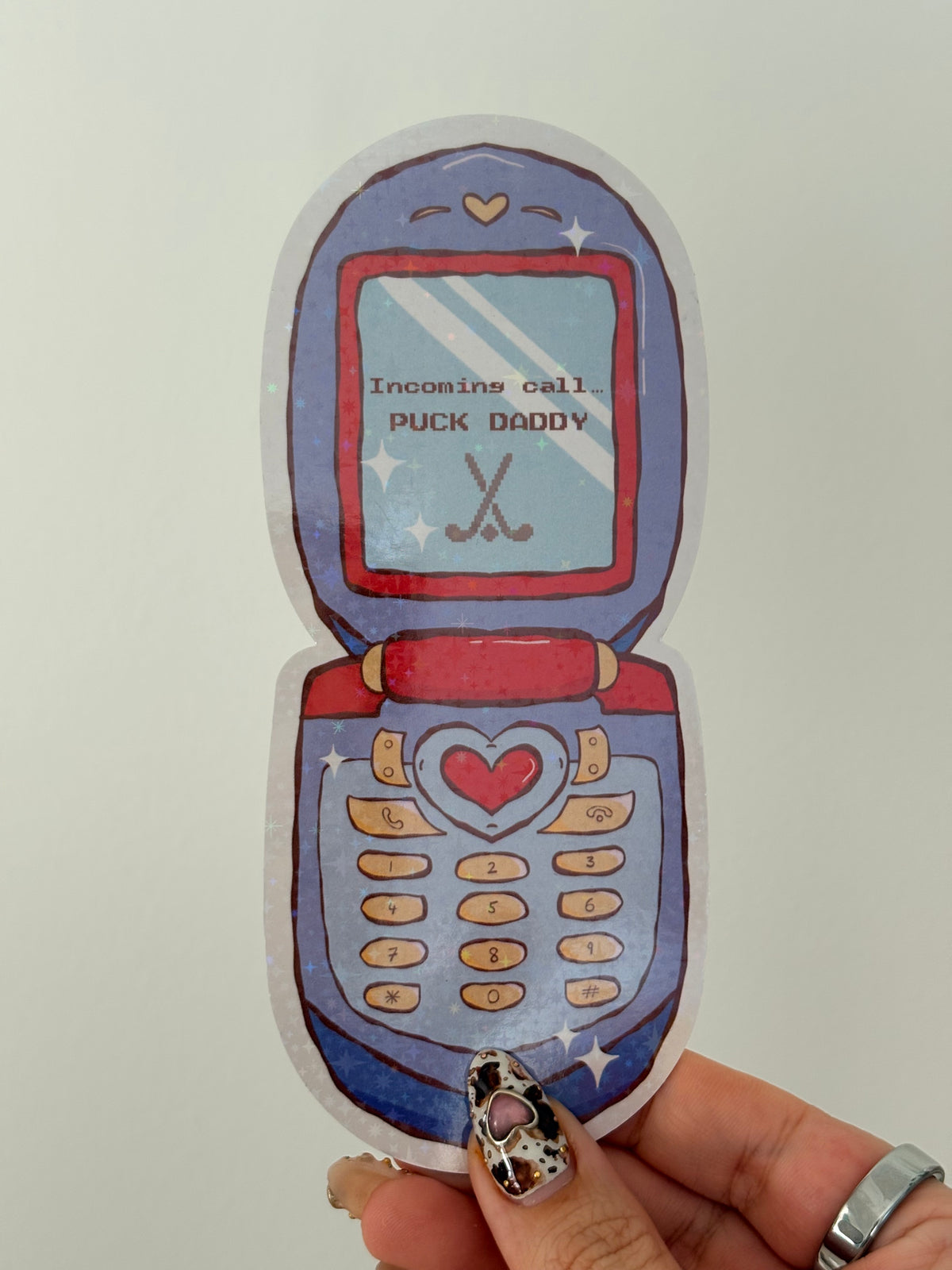 A novelty paper cutout bookmark shaped like a flip phone, held by a hand. The phone is light blue with red accents, gold buttons, and a screen that reads 'Innocent until PROUD DADDY'.