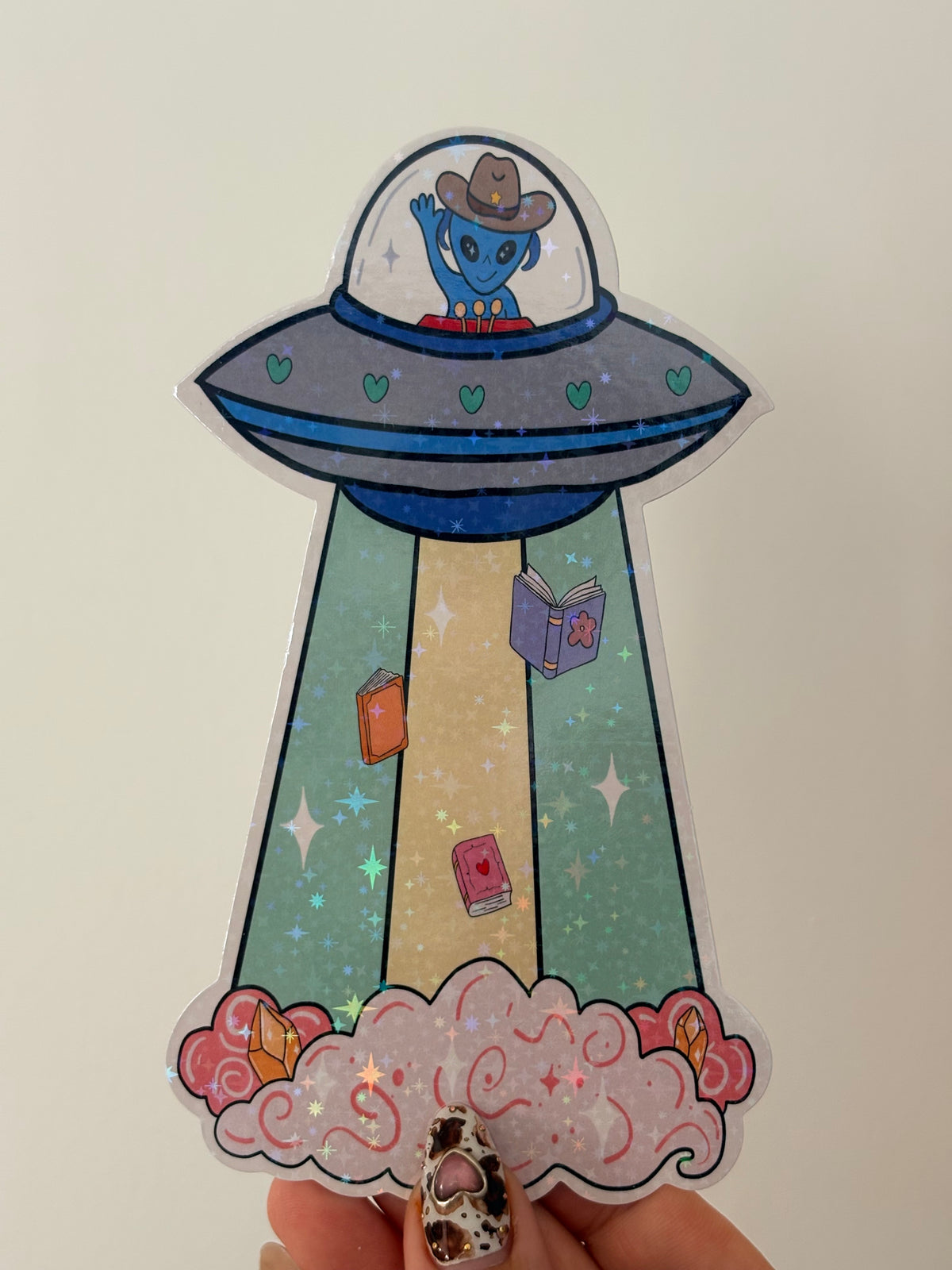 A hand holding a die-cut bookmark featuring a cartoon UFO with a blue character inside, a beige path with small colorful items, and pink clouds with glittery silver flecks.