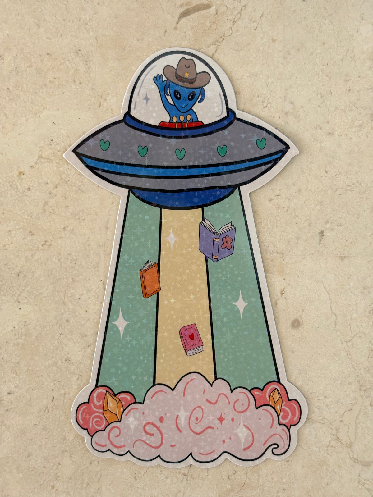 A die-cut bookmark featuring a cartoon UFO with a blue character in a cowboy hat inside, launching a beam of books into space with pink clouds at the end, shown on a textured beige background.