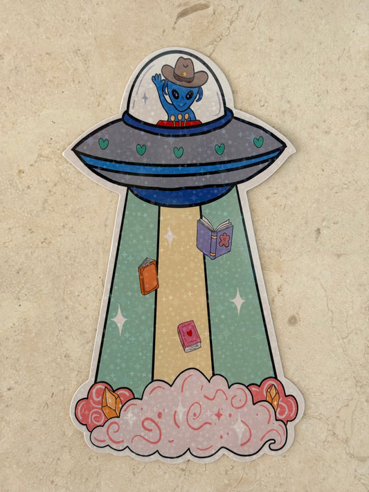 A die-cut bookmark featuring a cartoon UFO with a blue character in a cowboy hat inside, launching a beam of books into space with pink clouds at the end, shown on a textured beige background.