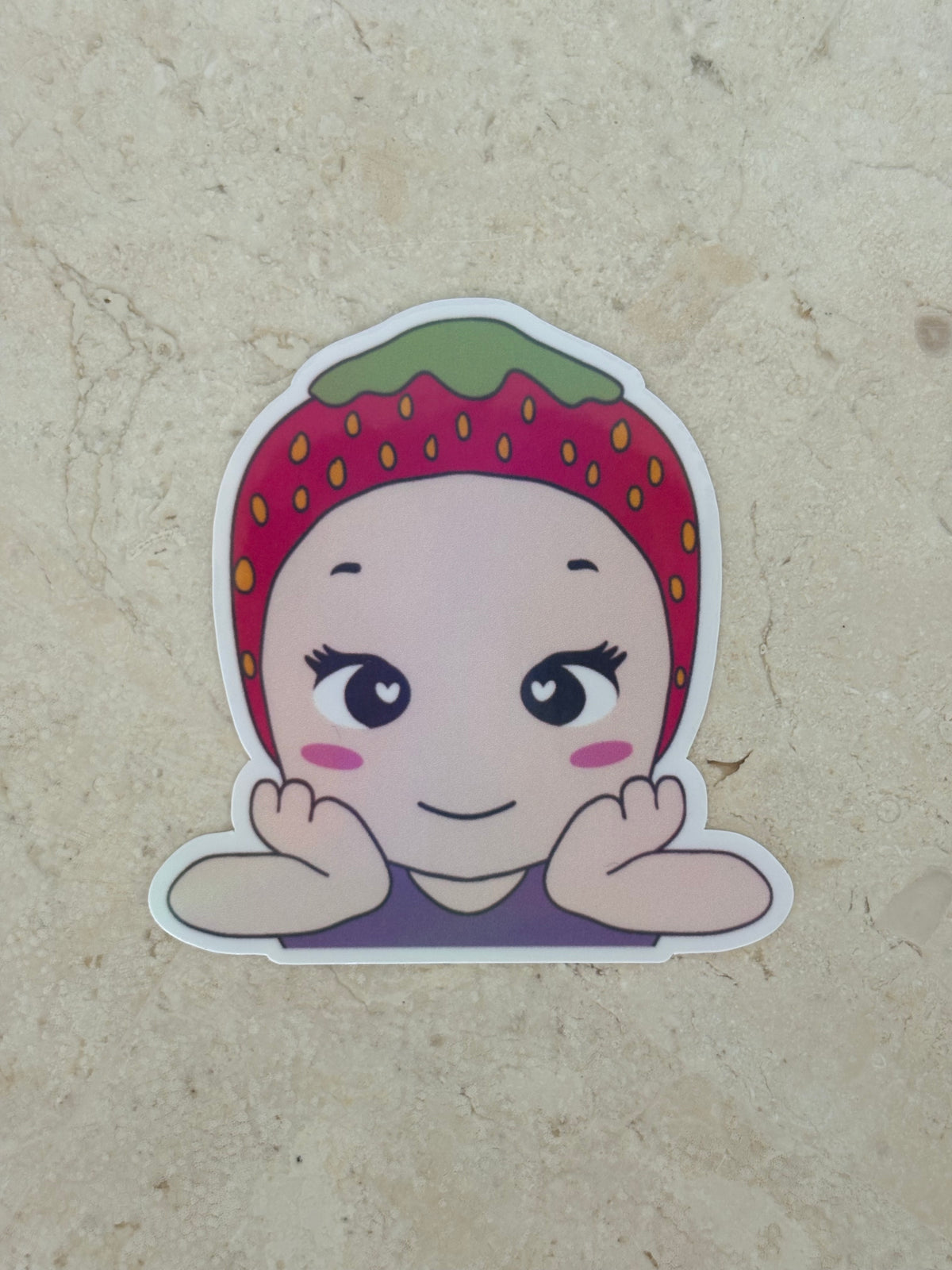 A die-cut sticker featuring a cute cartoon character with a strawberry-themed headband, resting on a beige textured surface.