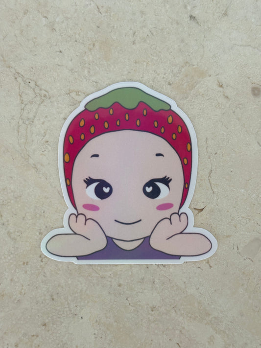 A die-cut sticker featuring a cute cartoon character with a strawberry-themed headband, resting on a beige textured surface.