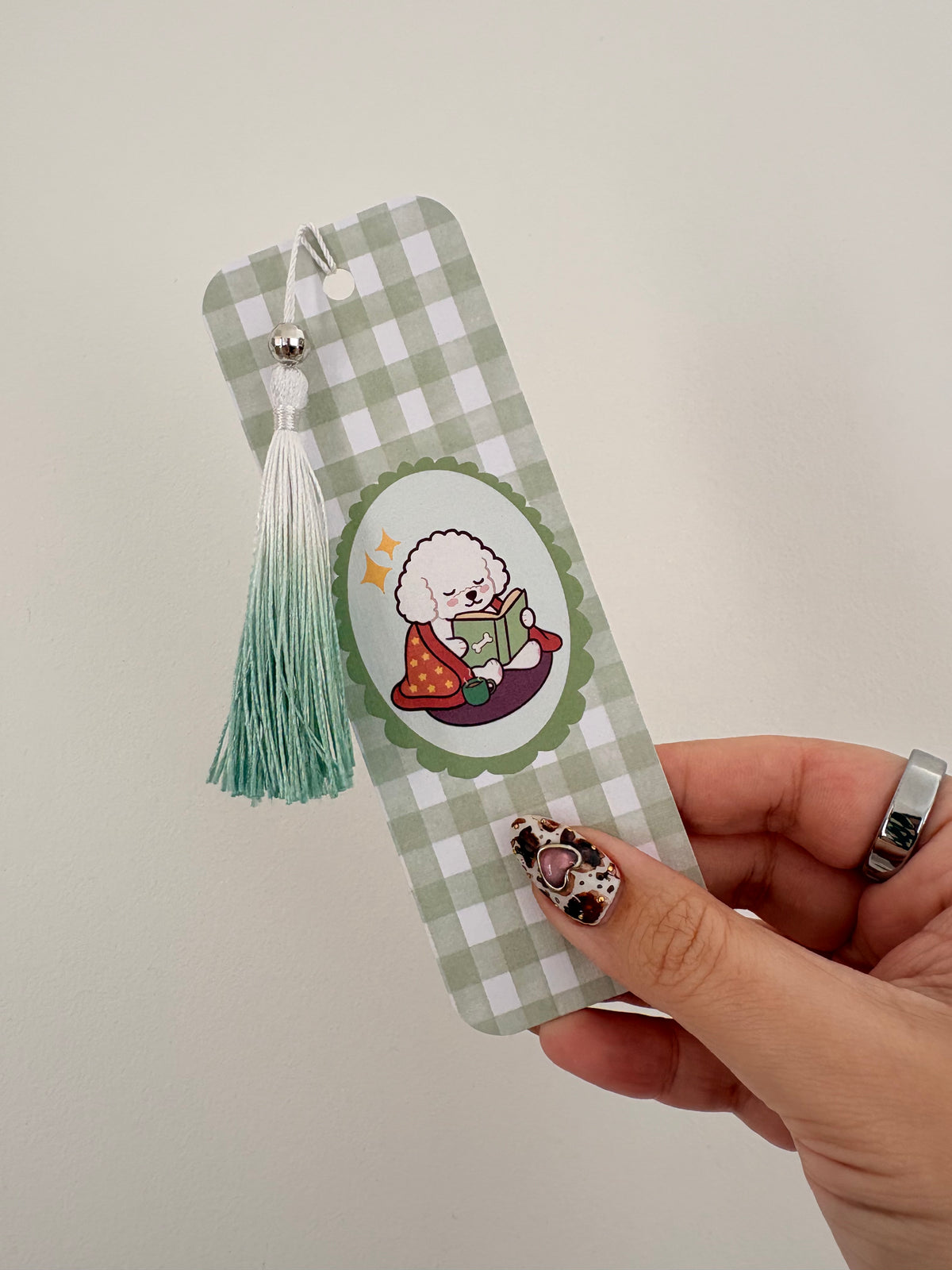 A hand holding a green and white gingham bookmark featuring a cartoon dog illustration and a mint green tassel, against a plain light background.