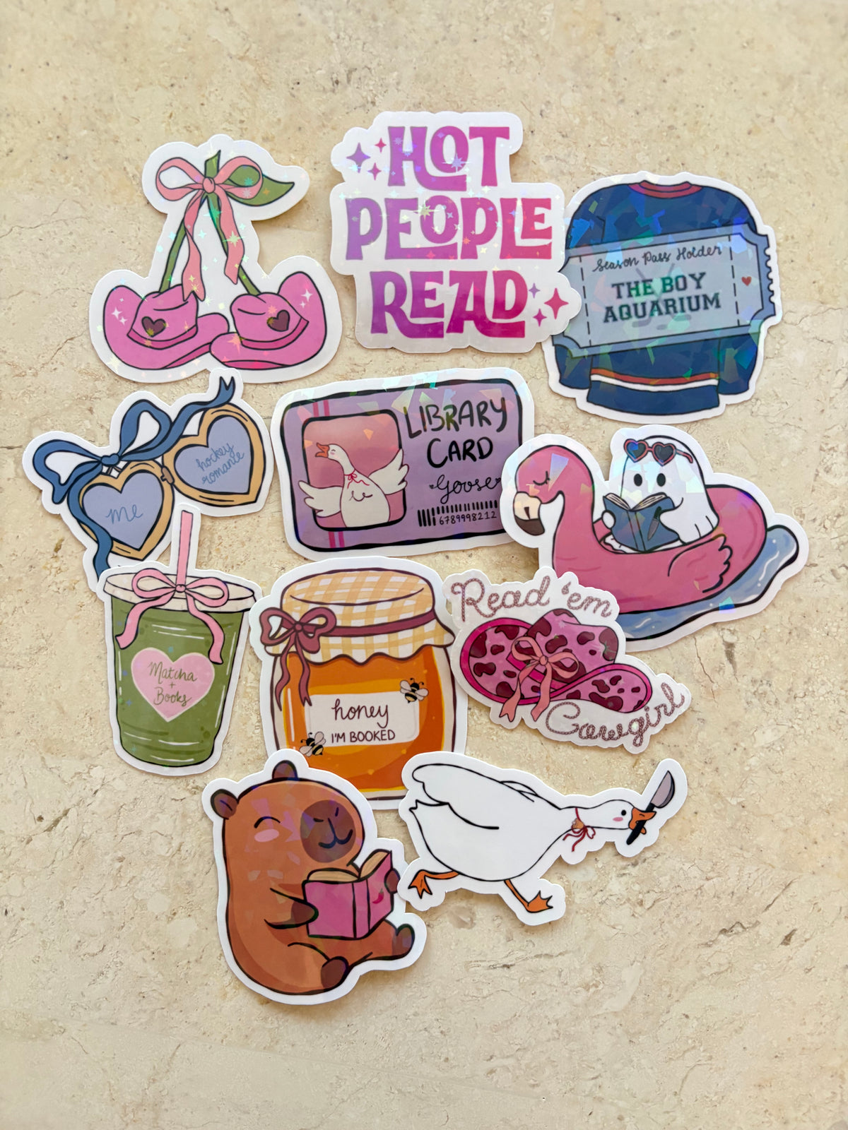 A set of ten die-cut sticker illustrations featuring cute, whimsical book-themed characters and objects, arranged on a textured beige surface. The stickers include a pair of pink ballet shoes, a blue sweater with a book label, heart-shaped sunglasses, a library card, a pink flamingo in an inflatable pool float, a green drink with a heart, a honey jar, a beaver reading, a white duck, and a goose with a book.