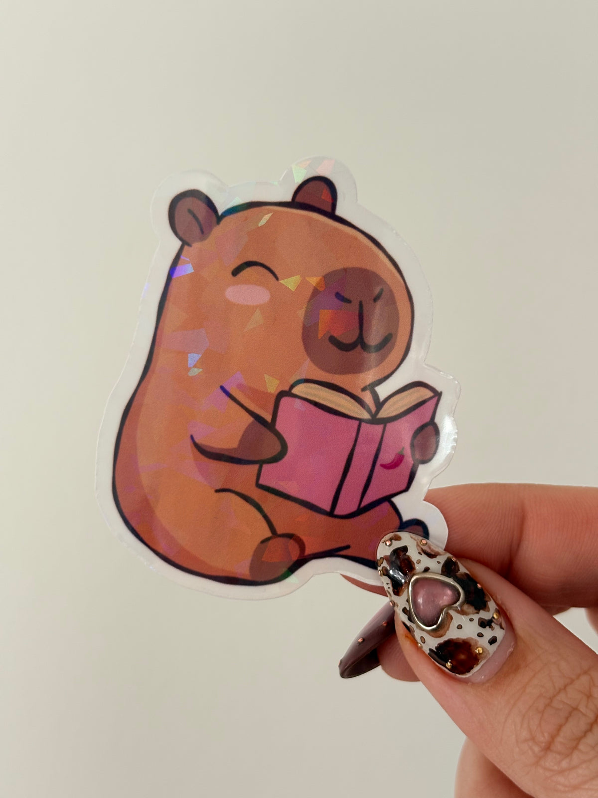 A die-cut sticker featuring a cute cartoon capybara holding a pink romance book.