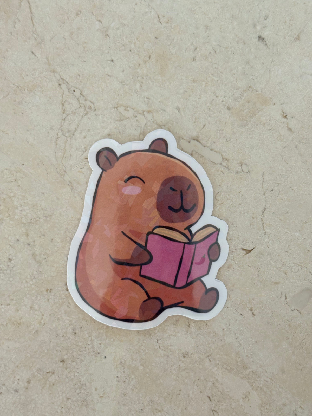 A die-cut sticker featuring a cute cartoon capybara holding a pink book.