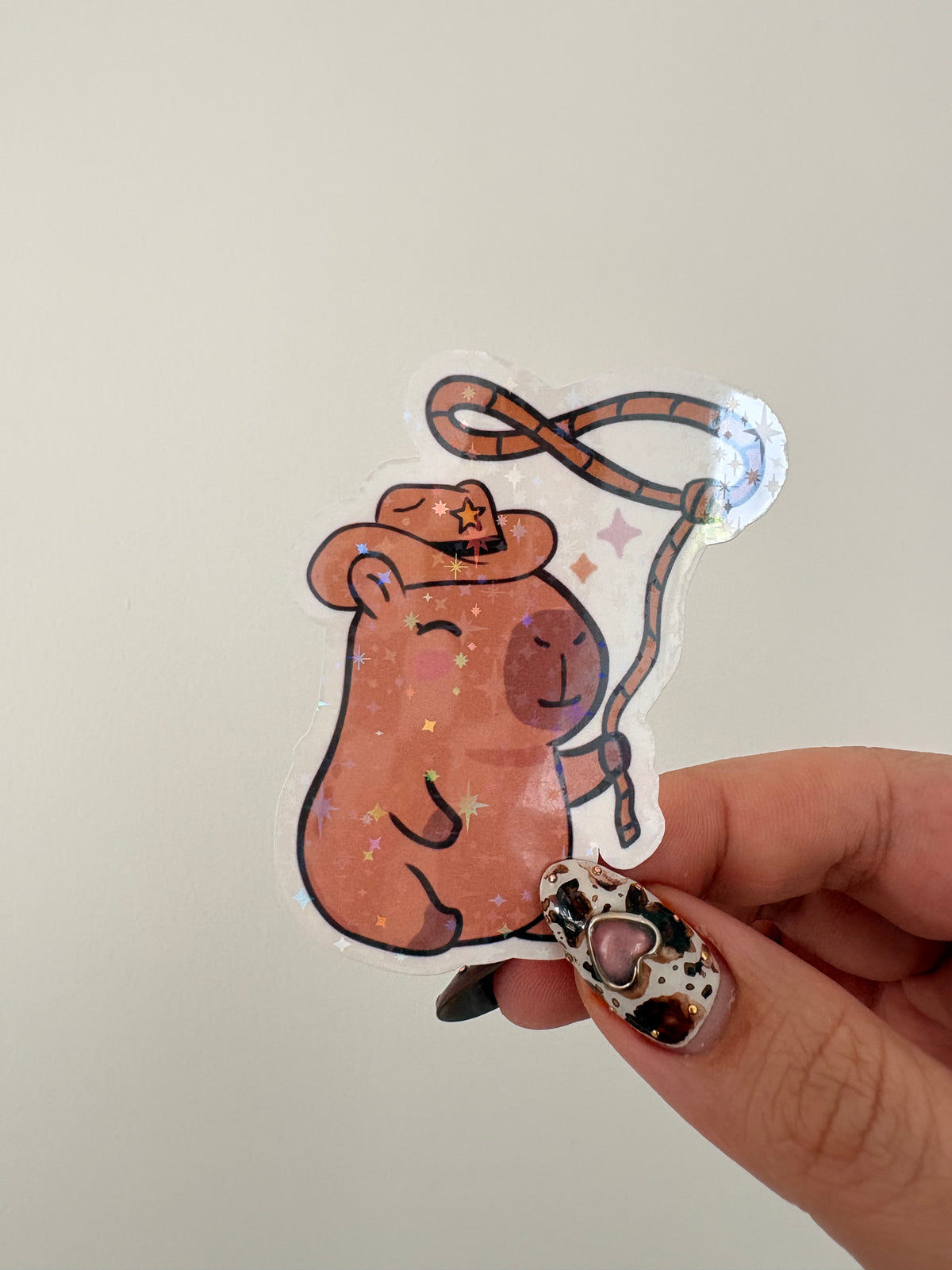 A hand with decorated nails holds a die-cut sticker featuring a cute brown capybara wearing a cowboy hat and holding a lasso, with a glittery finish, against a plain light background.