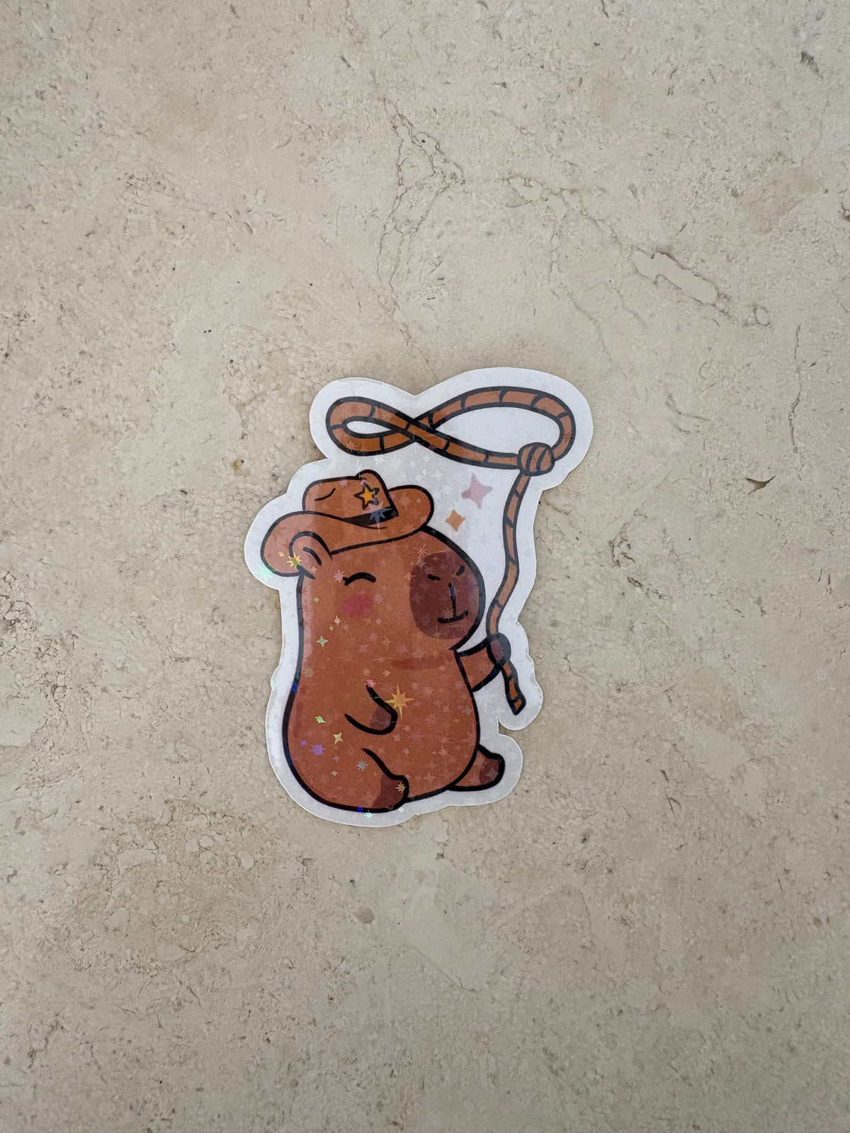 A die-cut sticker featuring a cute brown capybara wearing a cowboy hat and holding a lasso, placed on a textured beige stone surface.
