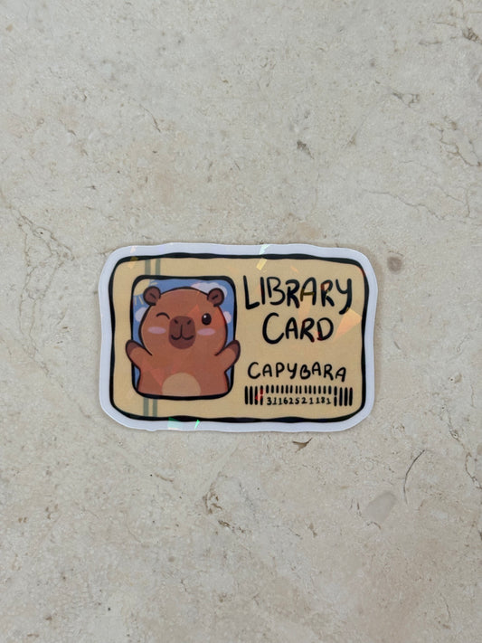 A die-cut sticker shaped like a library card featuring a cute cartoon capybara, placed on a textured beige stone surface. The sticker has a white border, a tan card background, and black text reading 'LIBRARY CARD' and 'CAPYBARA'.