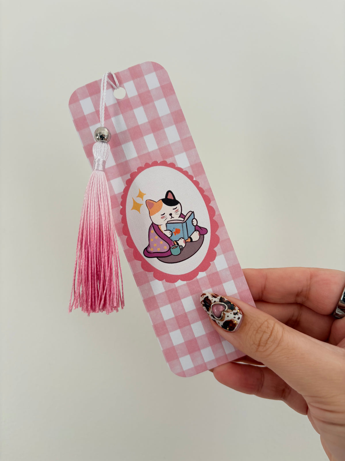 A pink and white gingham bookmark with a pink tassel and a cartoon cat illustration, held by a hand against a plain light background.