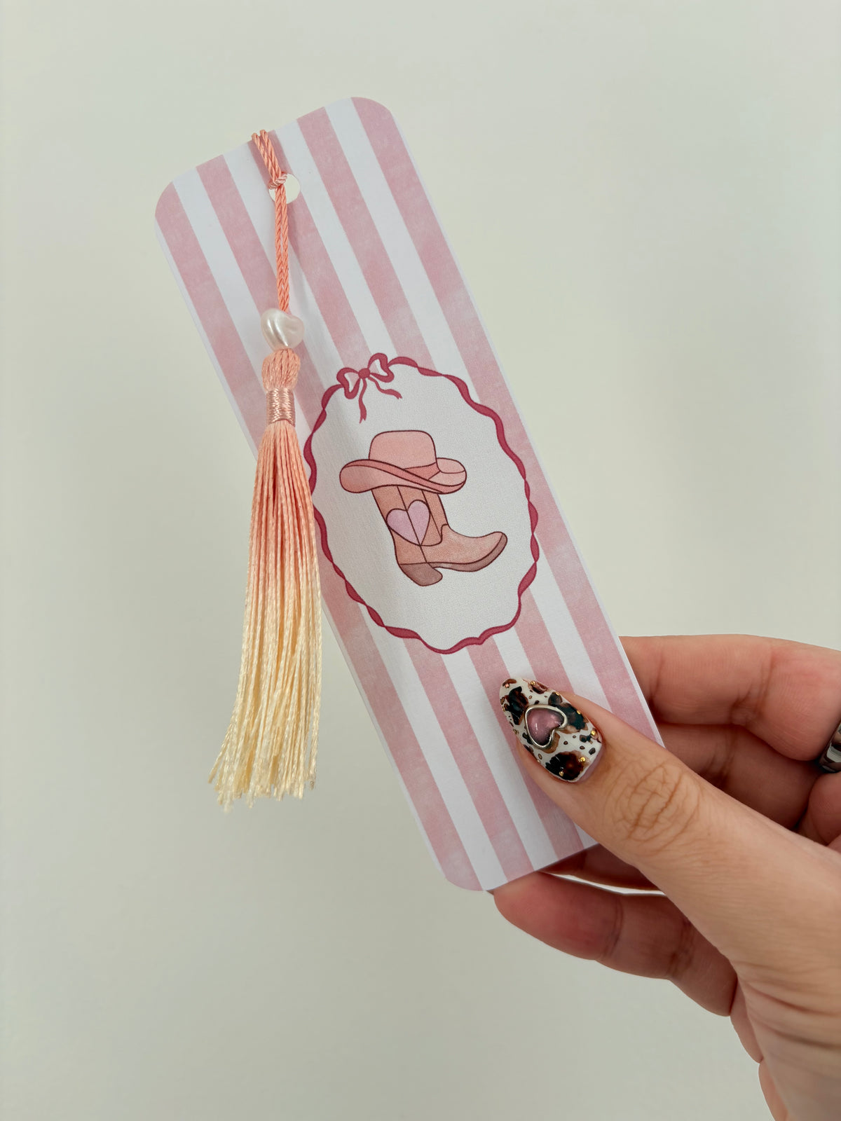 A hand with leopard-print nail art holds a pink and white striped bookmark featuring a cowboy boots illustration and a peach tassel, against a plain light background.