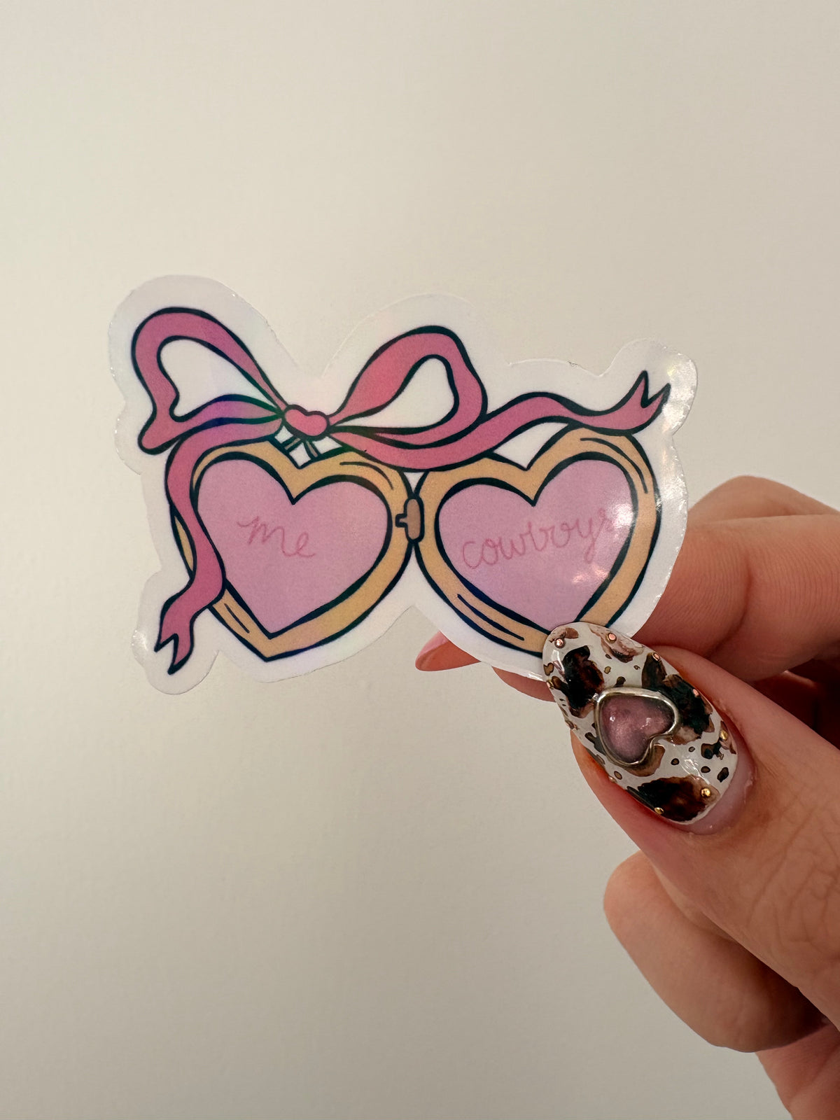 A hand with leopard-print nail art holds a die-cut sticker shaped like heart-shaped glasses with a pink ribbon bow. The sticker has pink lenses with the words 'me' and 'cowboy' written in cursive, outlined in gold, against a plain off-white background.