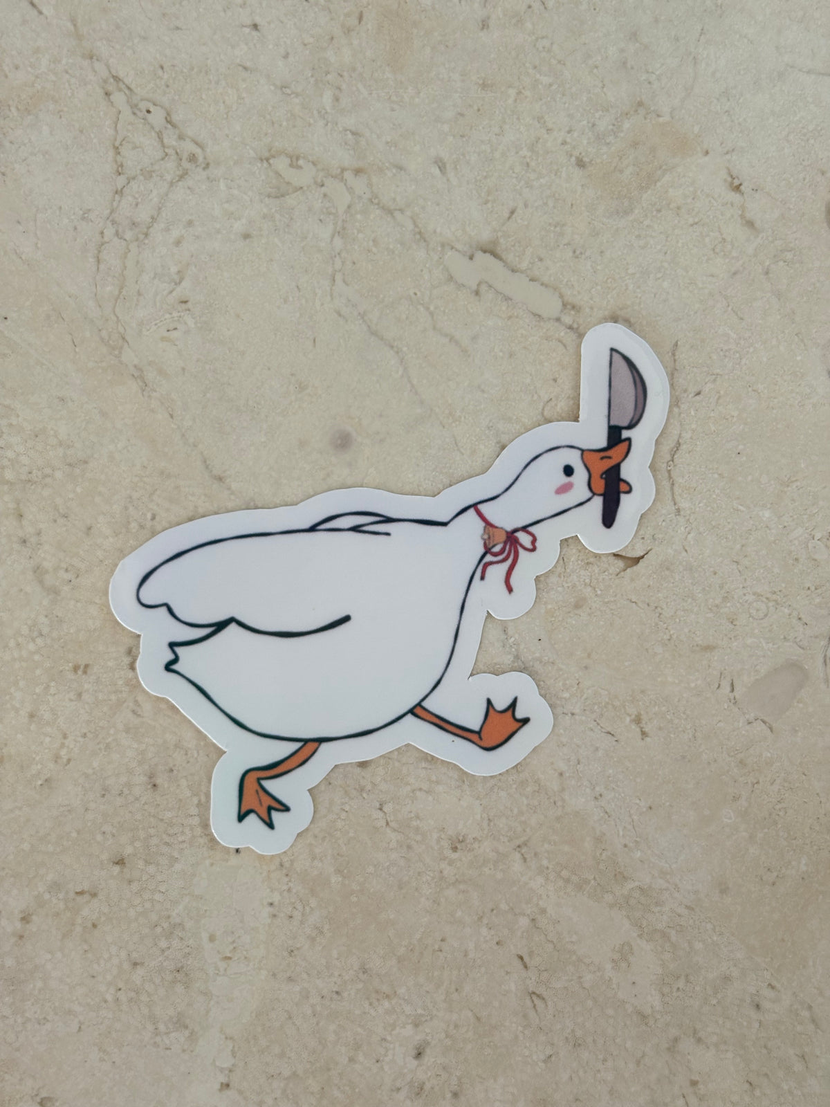 A die-cut vinyl sticker featuring a cartoon goose holding a knife and a pink bow tie, placed on a textured beige stone surface.