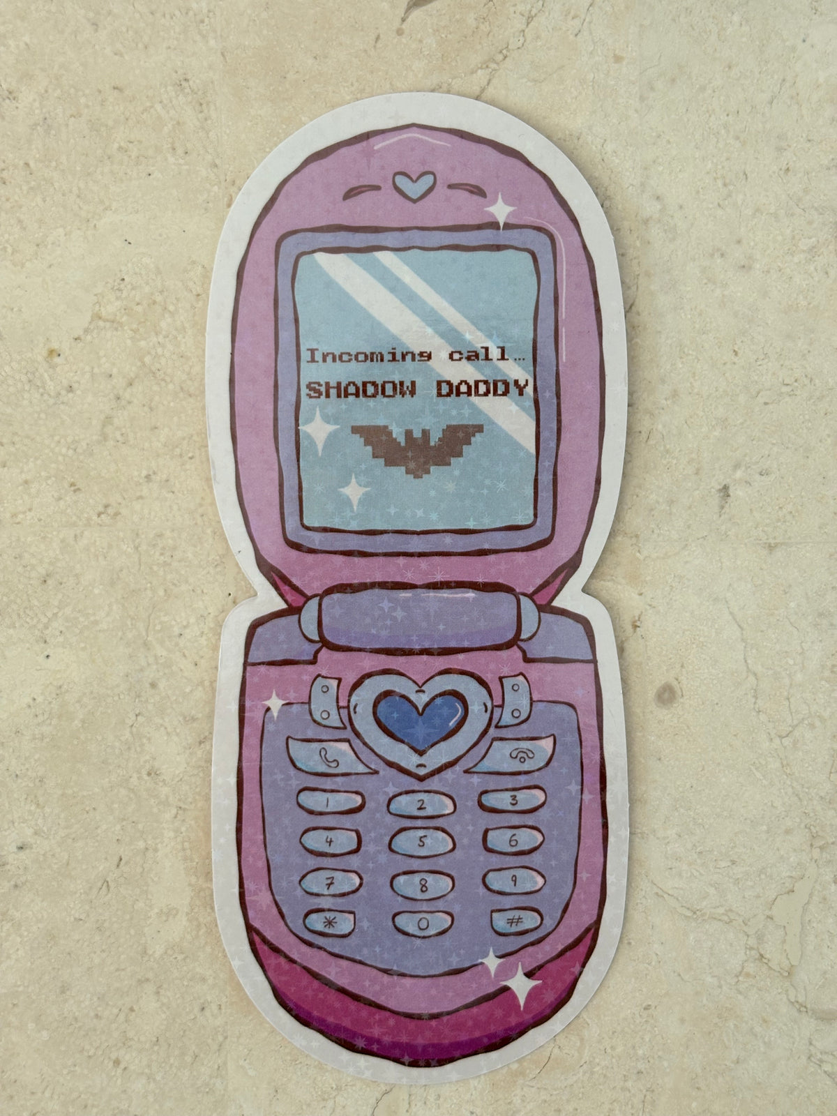 A die-cut sticker shaped like an open pink and purple flip phone, featuring a heart-shaped center button, star accents, and the text 'Incoming call: SHADOW DADDY' on the screen, displayed on a textured beige surface.
