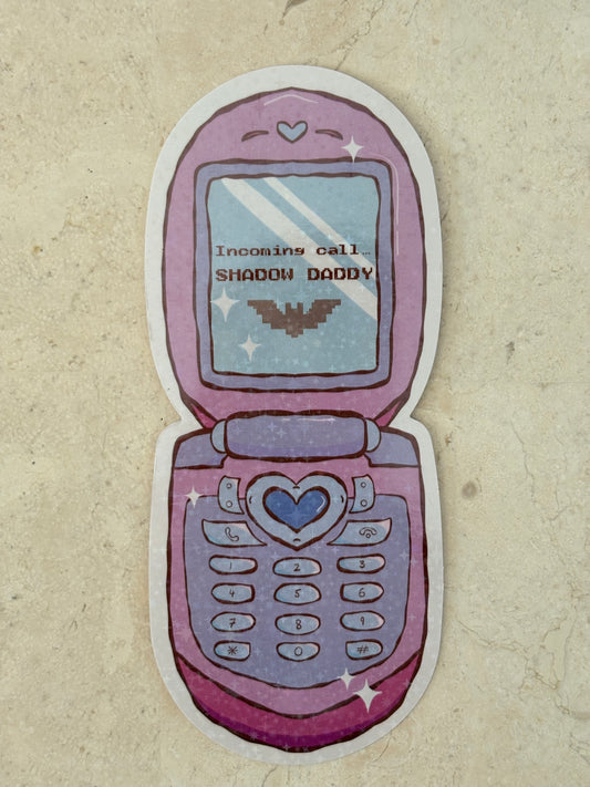 A die-cut sticker shaped like an open pink and purple flip phone, featuring a heart-shaped center button, star accents, and the text 'Incoming call: SHADOW DADDY' on the screen, displayed on a textured beige surface.