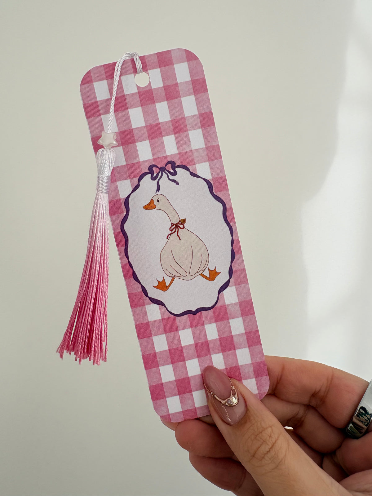 A pink and white gingham check bookmark held by a hand. The bookmark features a central white oval with a cartoon duck wearing a purple bow and orange beak/wings, and a pink tassel hanging from the top.