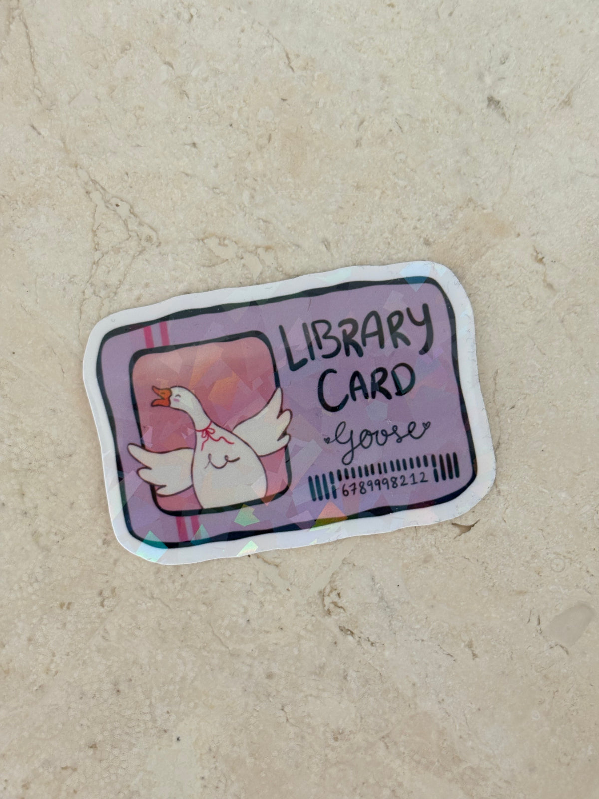 A die-cut sticker shaped like a library card featuring a cartoon goose, placed on a beige textured surface. The sticker reads 'LIBRARY CARD' with 'goose' written below, and includes a barcode and a pink-toned header.
