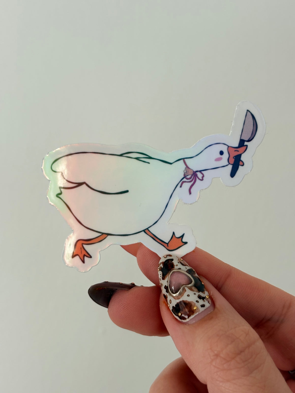 A die-cut sticker of a cartoon duck holding a knife, held by a hand with patterned nail art against a plain light background.
