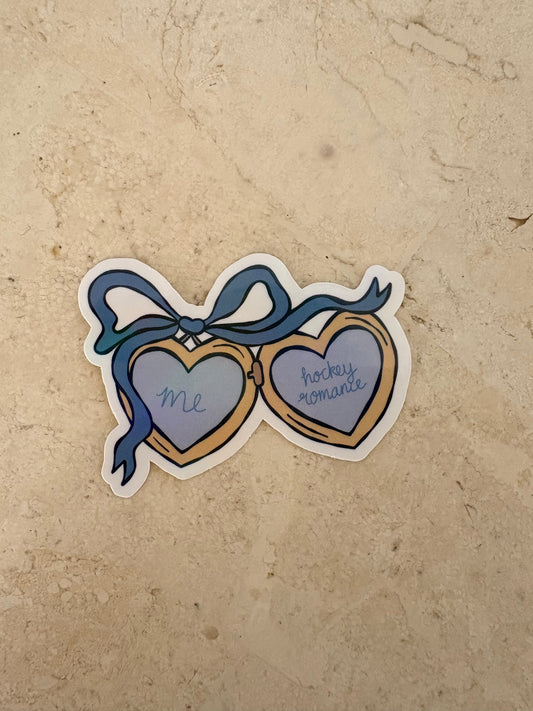 A die-cut sticker featuring a pair of heart-shaped sunglasses with a blue ribbon bow. One heart  with the word 'Me' in blue script, and the other is gold with 'hockey romance' in blue script.