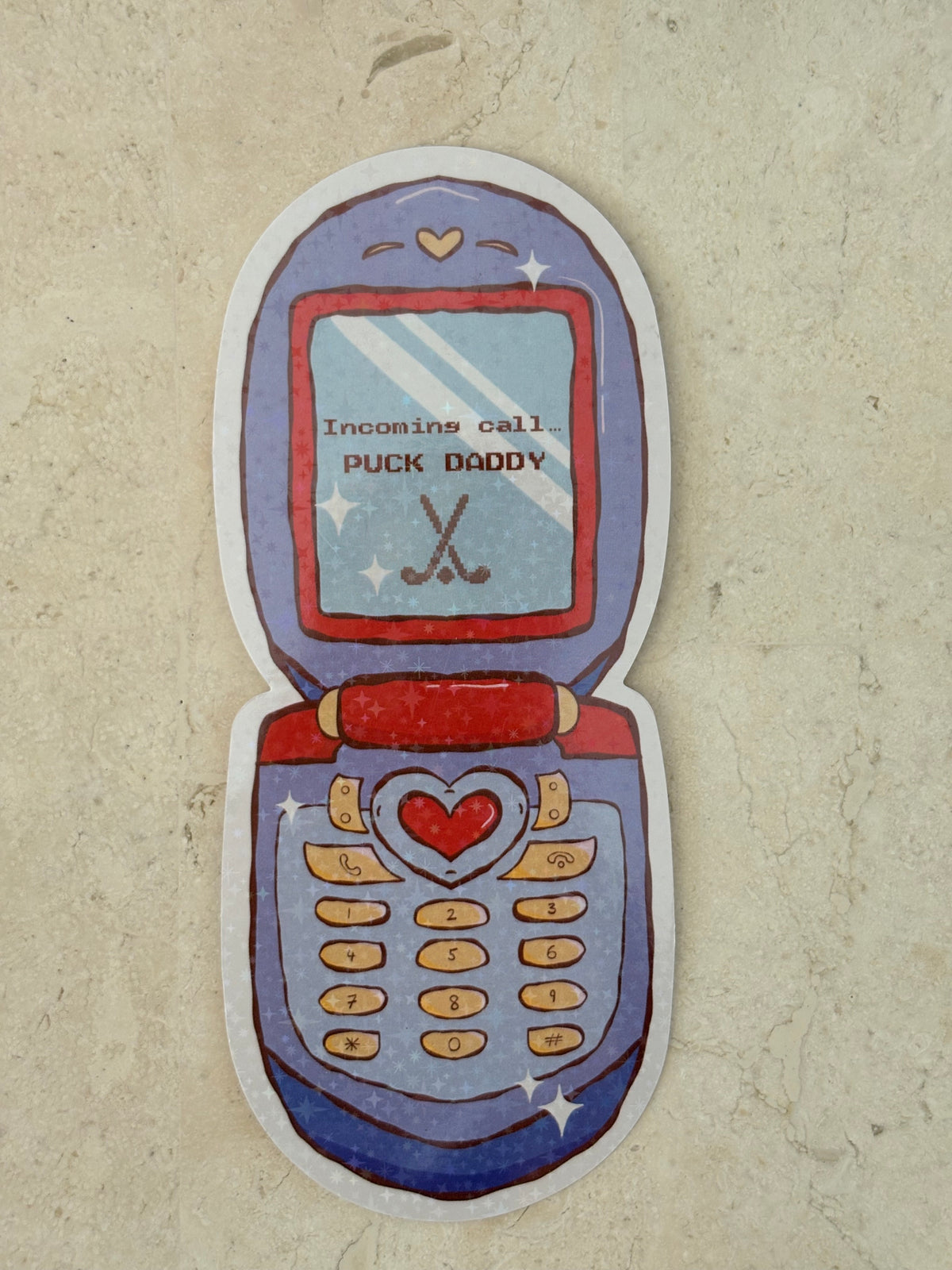 A novelty die-cut bookmark shaped like an open flip phone, featuring a hand-drawn design in shades of blue, purple, red, and beige, with the text 'I'm calling PUCK DADDY' on the screen, displayed on a textured beige surface.