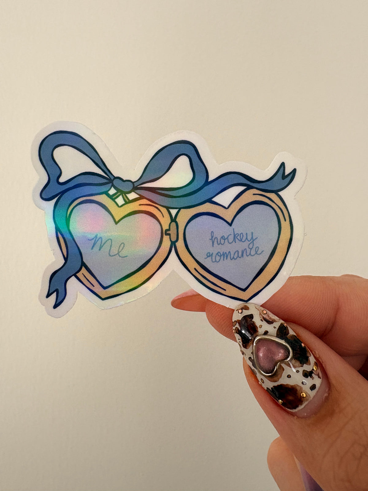 A die-cut sticker shaped like heart-shaped hockey goggles with a blue ribbon, held by a hand with decorated nails against a plain off-white background. The left heart says 'me' and the right heart says 'hockey romance' in blue script.