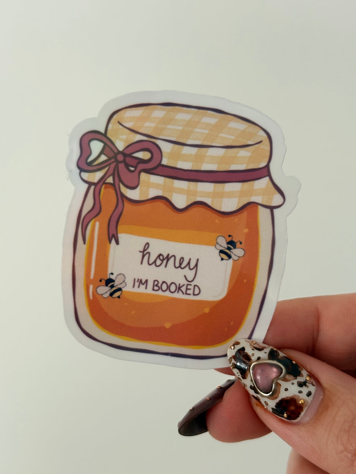 A die-cut sticker featuring an illustrated honey jar with the text 'honey I'M BOOKED' and small bumblebee details, held by a hand with patterned nail art against a plain light background.