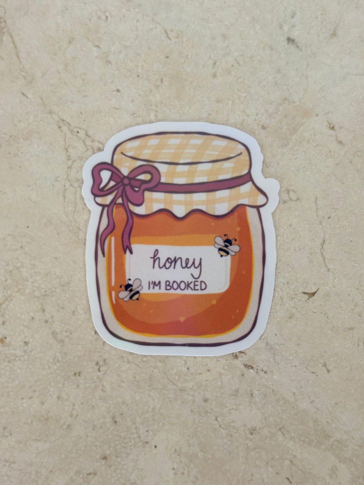 A die-cut sticker featuring an illustration of a honey jar with the text 'honey I'M BOOKED' and small bumblebee graphics, placed on a beige stone surface.