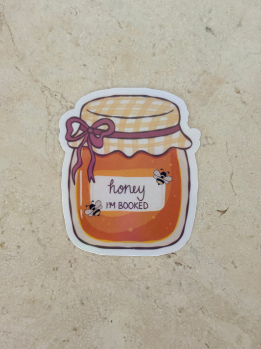 A die-cut sticker featuring an illustration of a honey jar with the text 'honey I'M BOOKED' and small bumblebee graphics, placed on a beige stone surface.