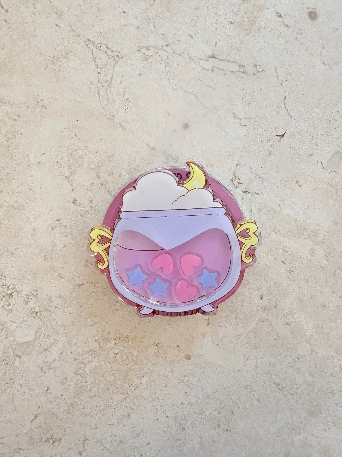 A cute acrylic charm of a purple cauldron filled with pink hearts and stars and a yellow moon.