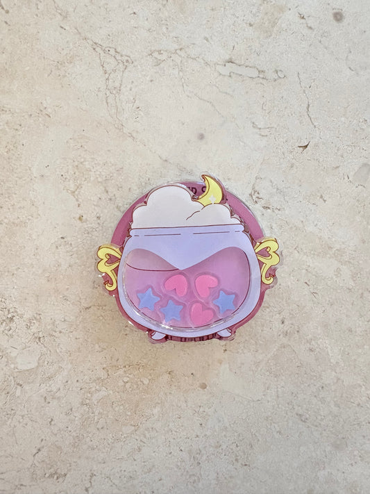 A cute acrylic charm of a purple cauldron filled with pink hearts and stars and a yellow moon.