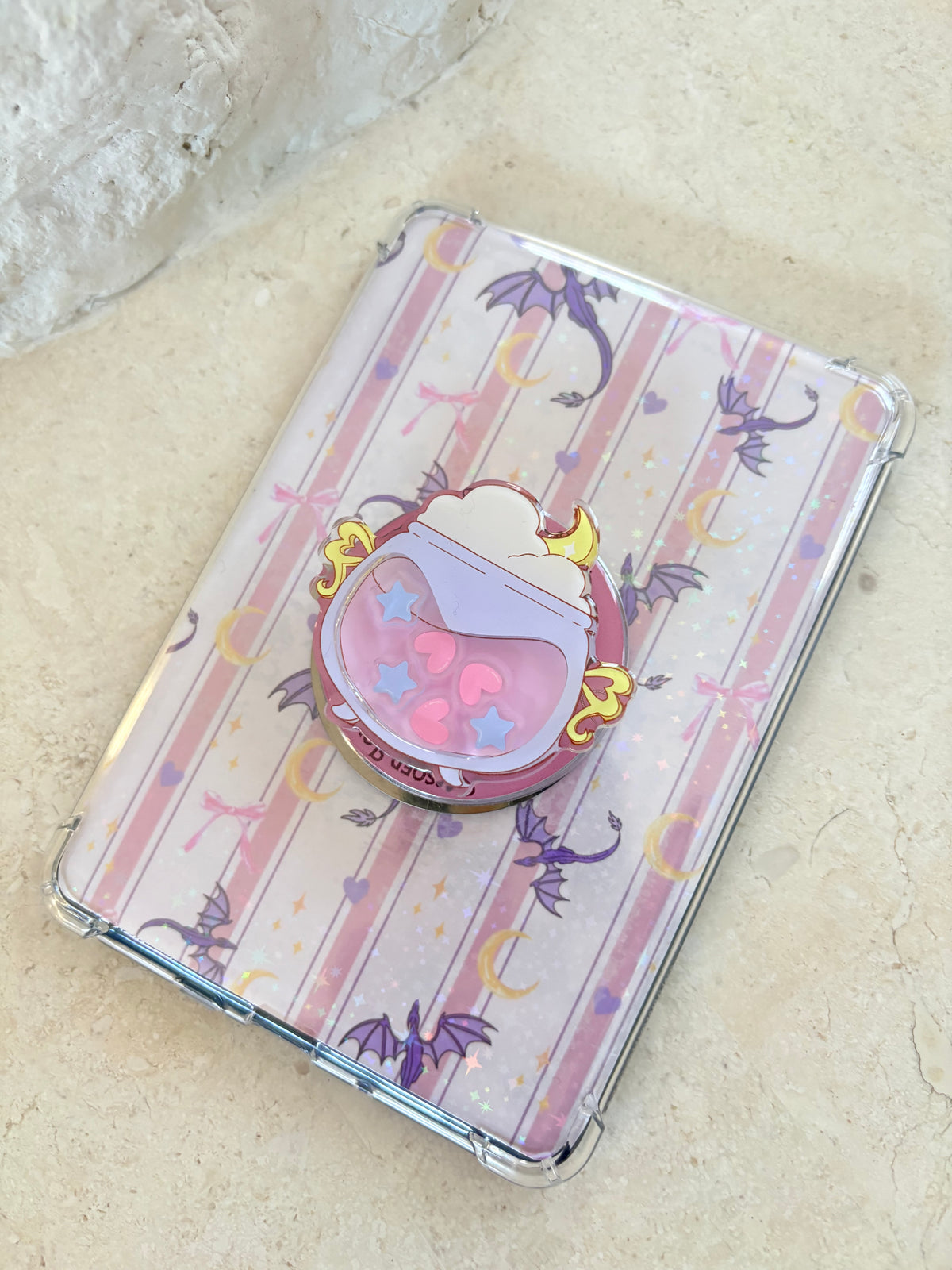 Cute cauldron enamel pin on a pink striped Kindle case with purple dragons.