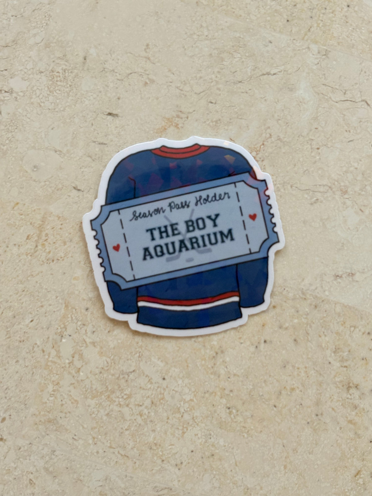 A die-cut sticker shaped like a hockey jersey ticket, featuring a navy blue jersey with red and white stripes and the text 'THE BOY AQUARIUM' and 'Season Pass Holder', placed on a textured beige surface.