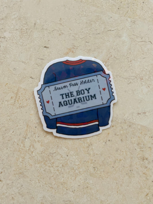A die-cut sticker shaped like a hockey jersey ticket, featuring a navy blue jersey with red and white stripes and the text 'THE BOY AQUARIUM' and 'Season Pass Holder', placed on a textured beige surface.