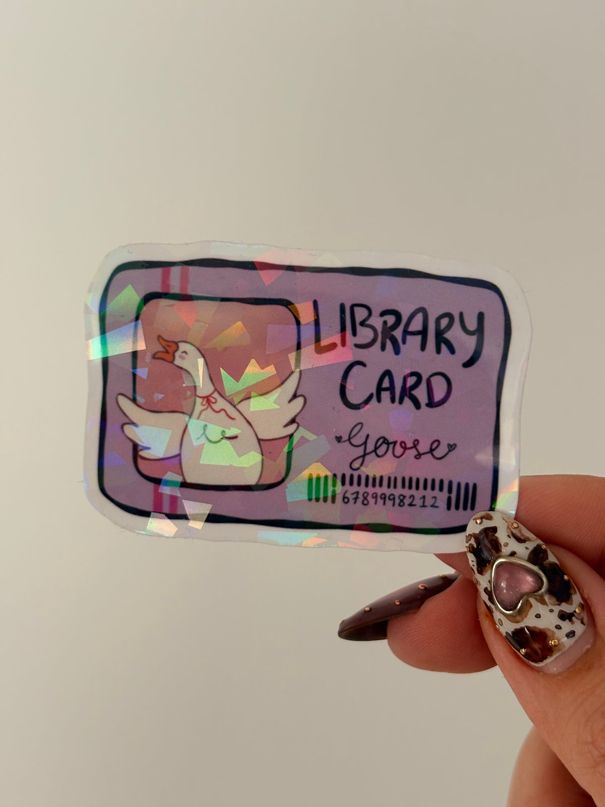 A hand with leopard-print nail art holds a holographic library card keychain featuring a cartoon goose and the text 'LIBRARY CARD Goose'.