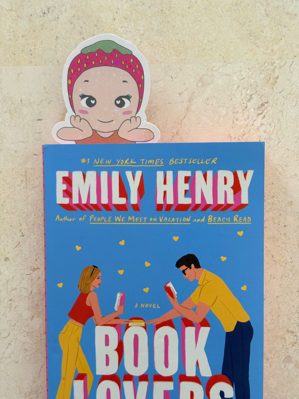 The book cover of 'Book Lovers' by Emily Henry, displayed on a textured beige wall with a cartoon sticker of a girl in a strawberry headband peeking out from behind the top edge of the book.