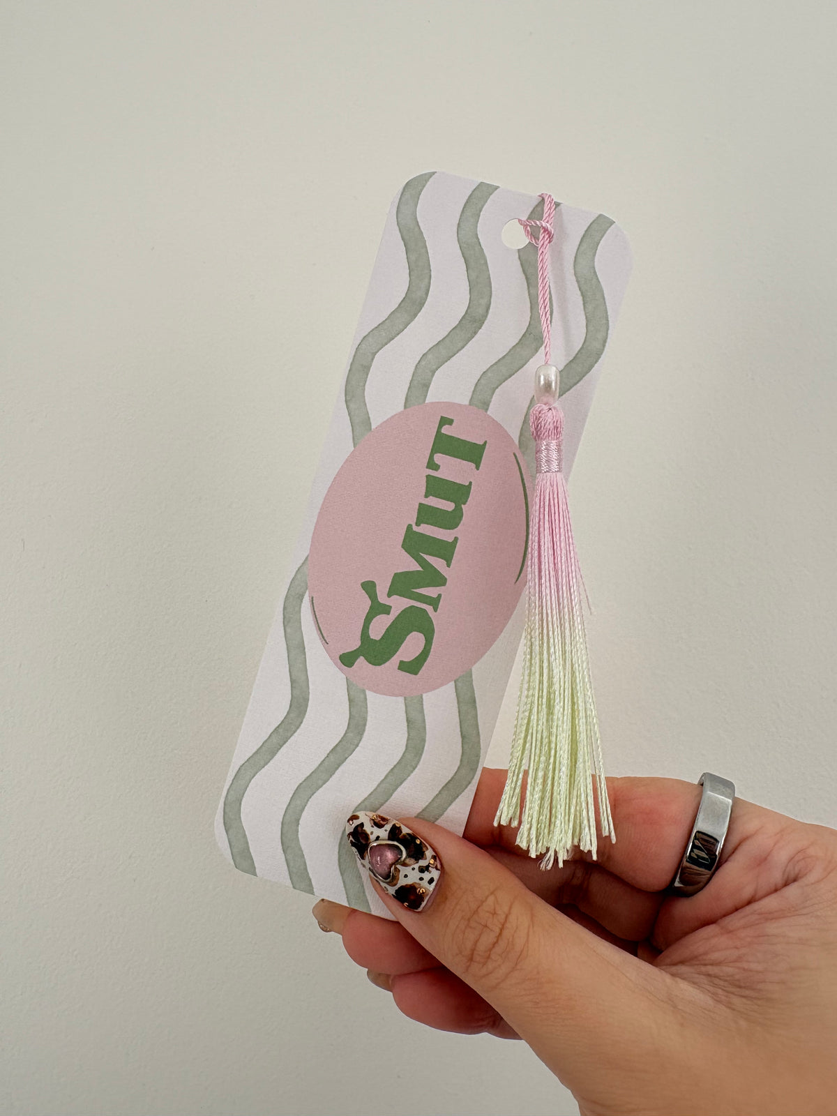 A hand holding a personalized paper bookmark with a pink tassel. The bookmark features a white background with wavy sage green stripes, a pink oval sticker with the name 'Smut' in green script, and a pink and cream tassel.
