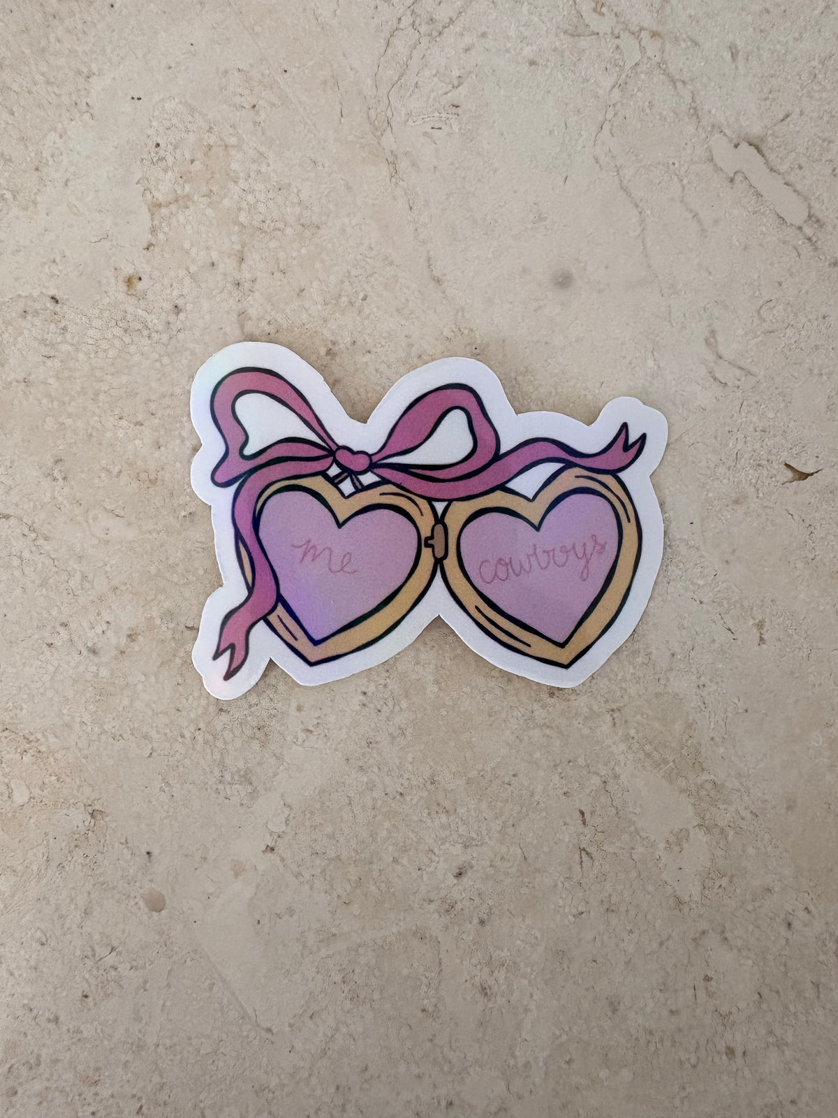 A die-cut sticker featuring a pair of heart-shaped sunglasses with pink ribbons, placed on a beige stone surface. The left heart is labeled 'me' and the right heart is labeled 'cowboys’