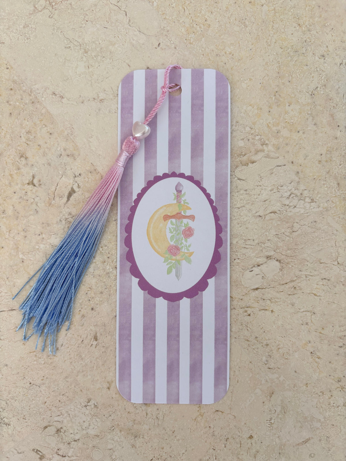 Purple and white striped bookmark with a crescent moon, sword, and pink-to-blue tassel.