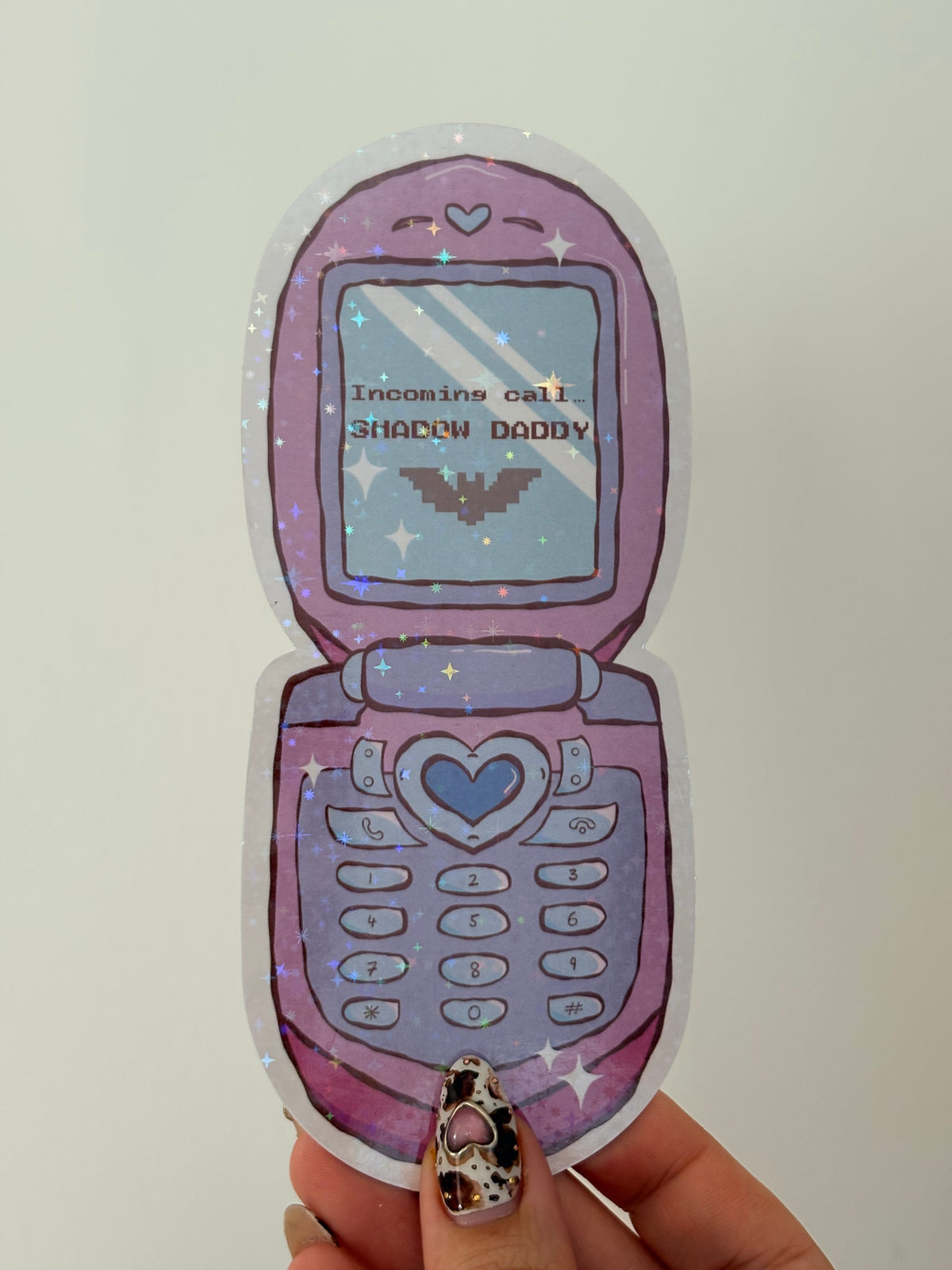 A hand holding a die-cut sticker shaped like a retro flip phone in pastel purple and lavender, with a heart-shaped center button, star details, and the text 'Ain't No Stupid Questions' on the screen.