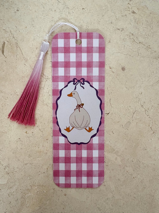 A pink and white gingham bookmark with a hanging tassel and a central illustration of a duck inside an oval frame, placed on a beige textured surface.