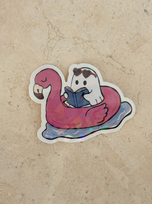 A die-cut sticker featuring a cute cartoon ghost wearing heart-shaped sunglasses and reading a blue book while floating on a pink flamingo float, placed on a beige stone surface.
