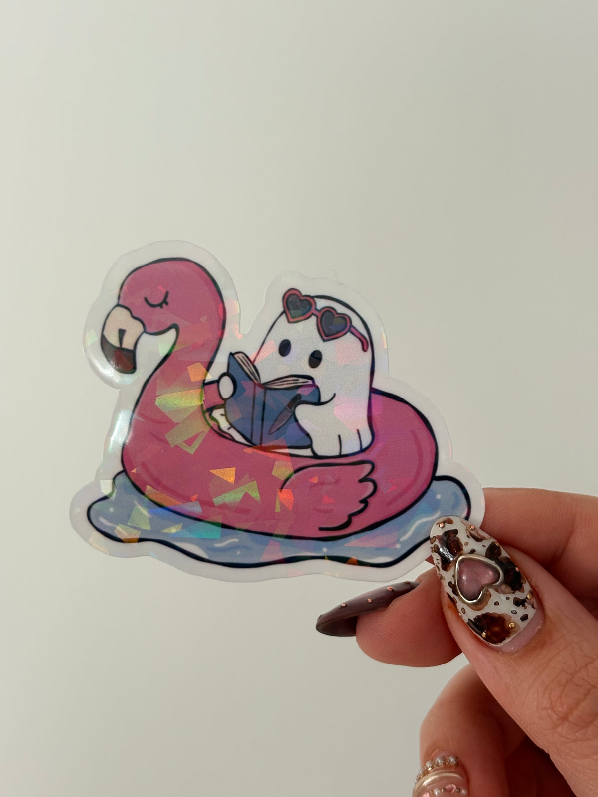 A hand with leopard-print nail art holds a die-cut acrylic charm featuring a white cat wearing sunglasses and hugging a pink flamingo-shaped floatation ring with holographic accents, set against a plain light background.