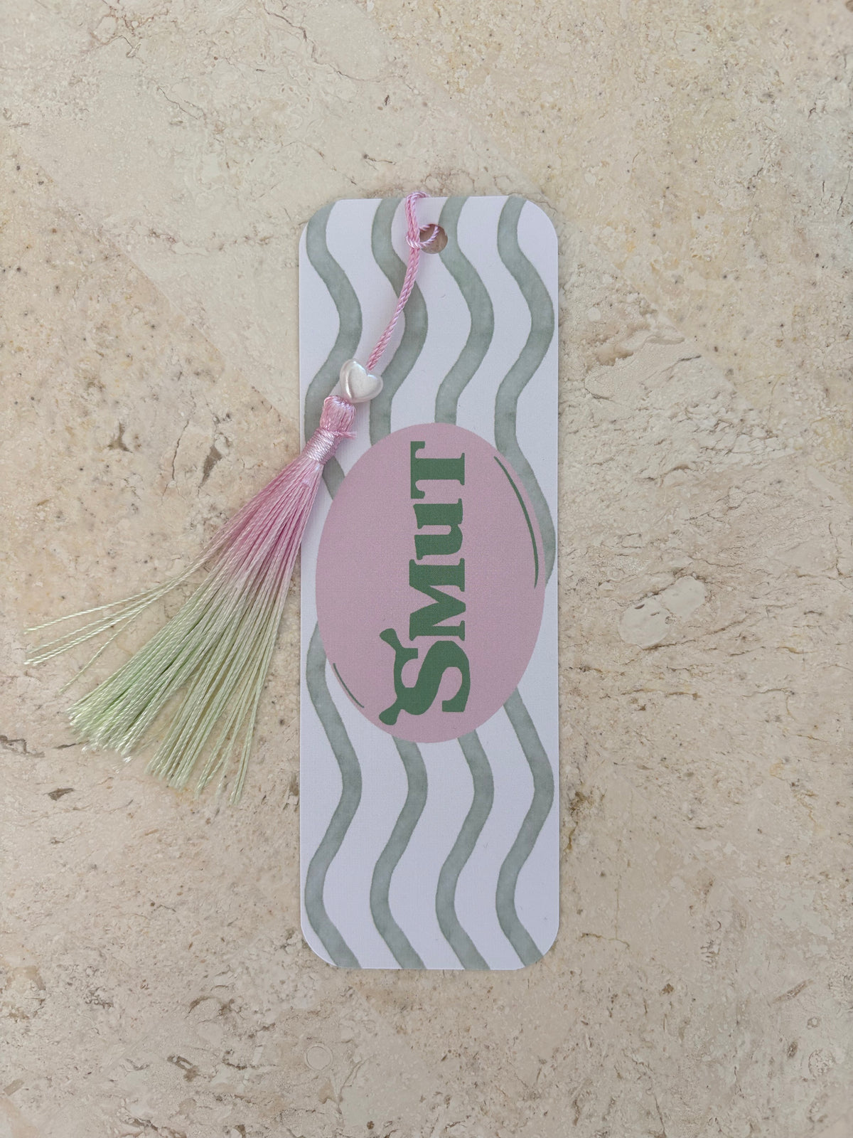White bookish bookmark with grey waves, pink oval and 'SMUT' text, with a pink and green tassel.