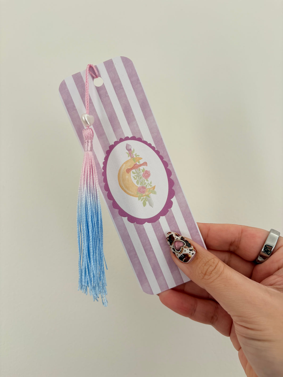 A hand holding a lavender and white striped bookmark featuring a central oval illustration of a crescent moon, flowers, with a pink and blue gradient tassel hanging from the top.